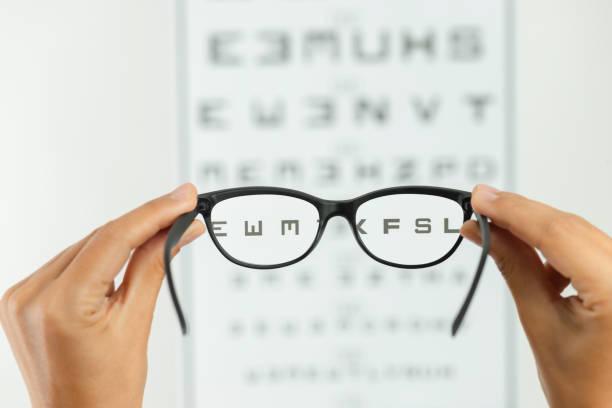 Eye Exam Hand is holding reading glasses in front of an eye exam chart in front of a white wall. reading glasses stock pictures, royalty-free photos & images
