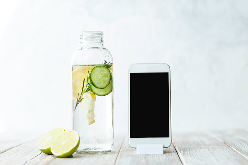 Bottle of infused water on white wood with a slice of lemon , cucumber and rosemary leaf in it. Smart phone next to the bottle. Representing detox blogging or mobile applications about detox and healthy lifestyle concept.