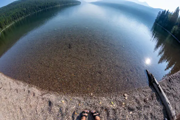 Fisheye wide angle view of Bowman Lake in Glacier National Park, with woman's feet with sandals on the shoreline