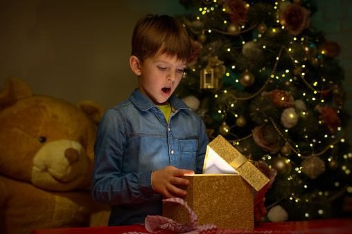 A surprised boy open big lightning box of New Year’s gift in front of a Christmas tree