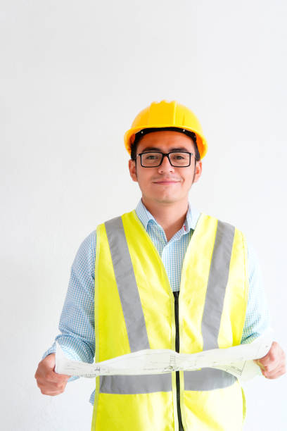 istock Young engineer looking at the camera holding a blueprint and carrying construction safety equipment, on white background with copy space. 1342849326
