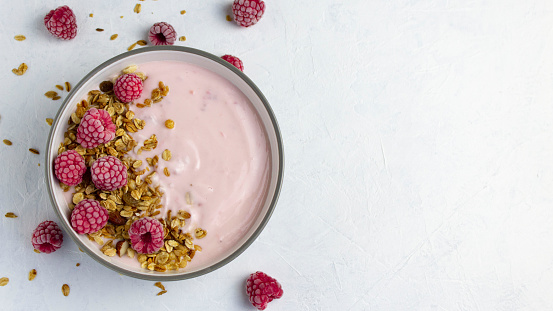Top view of raspberry yogurt  bowl with granola and frozen raspberries on a white plate with a copy space