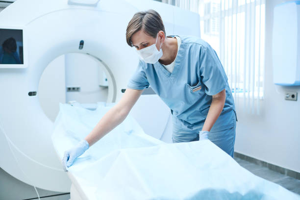 istock Short-haired nurse in surgical mask and gloves spreading out disposable sheet on table while preparing MRI scan for patient 1342881856