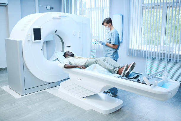 MRI specialist in facial mask and gloves standing at patient lying on table and filling his card online MRI specialist talking to patient cat scan stock pictures, royalty-free photos & images