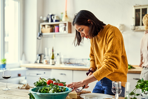 Young woman chopping vegetables on kitchen island with her friend in background. Woman making food at home.