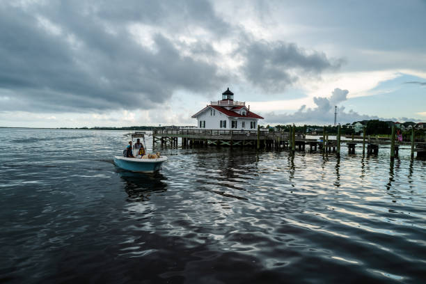 A scenic view of a wooden pier with green pilings, overlooking a lighthouse with a red roof located on the waterfront.