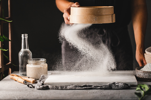 Cropped shot of a woman making bread in the kitchen. Hand of a female sifting the flour with an old wooden sieve onto a marble board to make sourdough bread dough.