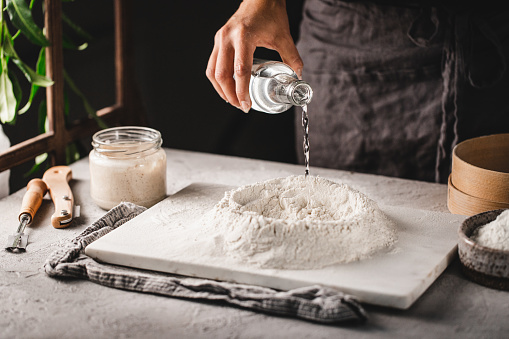 Cropped shot of woman pouring water on rye and wheat flour over marble board. Female in the kitchen preparing sourdough bread dough.