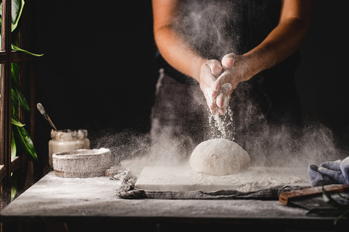 Close-up of woman kneading bread dough on marble board over kitchen counter. Midsection of female preparing sourdough bread in kitchen sprinkling flour over the dough.