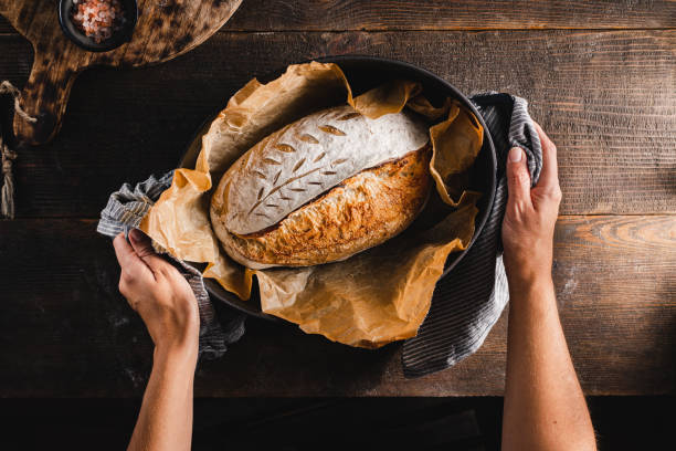 Woman baking sourdough bread in kitchen Point of view of a female with a freshly baked loaf of sourdough bread in a baking tray. Woman making sourdough bread in the kitchen. baking bread stock pictures, royalty-free photos & images