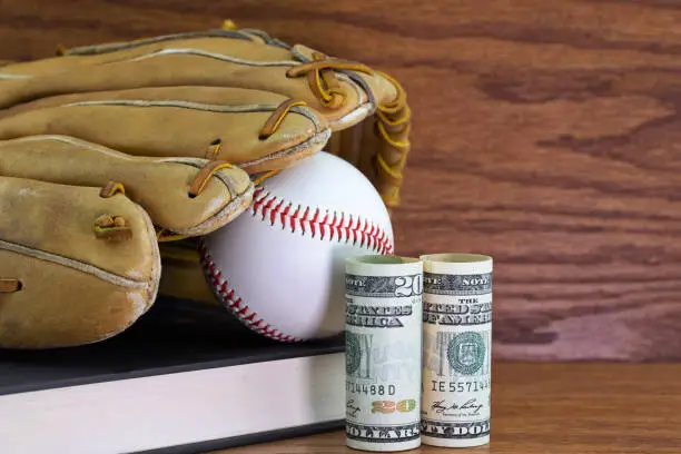 Scholar athlete concept symbolized in dollars placed with black textbook; baseball glove and ball on formal wood background