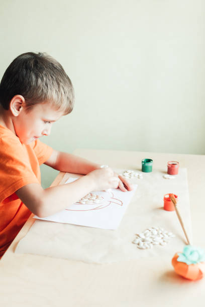 menino de 7 anos fazendo abóbora de sementes em papel branco sentado à mesa - child autumn craft preschooler - fotografias e filmes do acervo