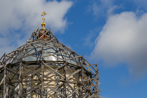 gray wood scaffolds on a dome of russian christian church with golden religious cross at day light on blue sky with white clouds