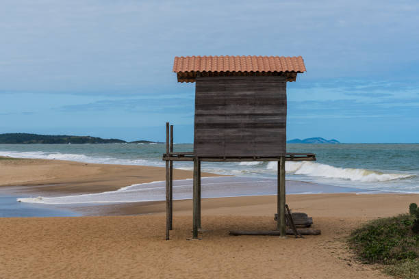 estrutura de madeira em forma de salva-vidas na praia de rio das ostras, no rio de janeiro. dia parcialmente nublado, céu azul e algumas nuvens. mar forte e areia amarelada e muitas rochas - villa summer rock sand - fotografias e filmes do acervo