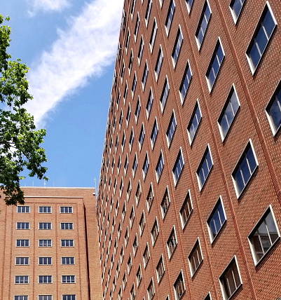 Looking up at a tall brick residential dormitory apartment structure. Building has many three panel glass windows. Classic architecture style in an urban midwest or east coast United States city. Blue sky above with a sweeping white cloud in sight. Glimpse of green leaves on a tree branch in upper left corner. Symmetry and angles  give the photo a mesmerizing energy and flow feel. Daytime shot. No people.