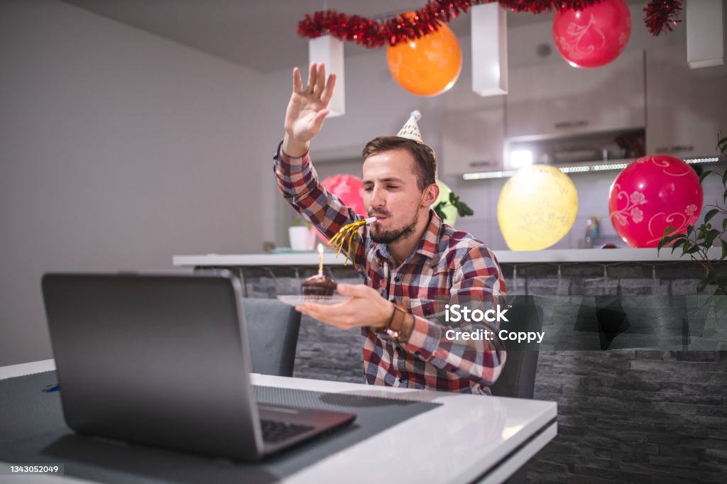 Young man celebrating his birthday with his friends on video call during pandemic COVID-19 coronavirus bday whistle Young man blowing birthday whistle with his friends on video call on laptop during pandemic COVID-19 coronavirus isolation 25-29 Years Stock Photo