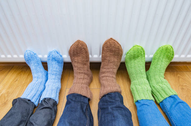 People wearing knitted warm wool socks warming cold feet in front of heater at cold home. stock photo