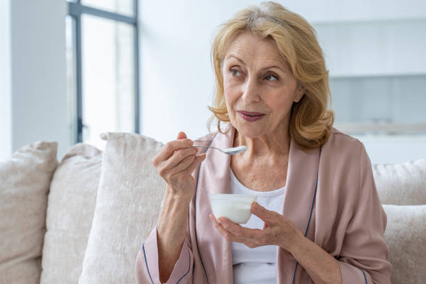 Smiling elderly woman who eats yogurt sitting on the couch stock photo