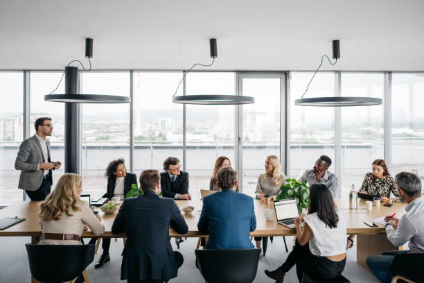 Business meeting in a bright office A photo of a business meeting in a modern office with large windows. A businessman is standing up while his colleagues are sitting down. They are smartly dressed. Horizontal daylight indoor photo. director stock pictures, royalty-free photos & images
