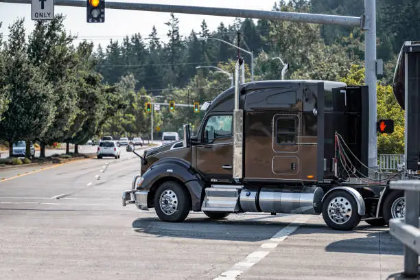 Photo of Black stylish compact cab big rig semi truck tractor transporting cargo in tented semi trailer turning on the city street crossroad with traffic light