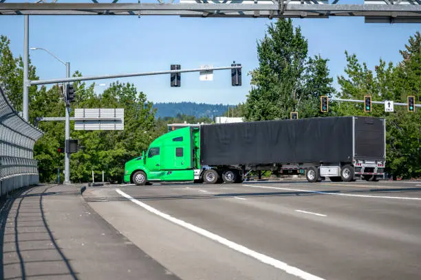 Photo of Big rig green industrial grade semi truck with black covered semi trailer turning on the city crossroad with traffic lights