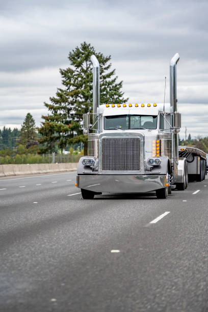 istock Classic light white big rig semi truck with tall exhaust pipes transporting empty flat bed semi trailer driving on the wide one way highway road 1343098709