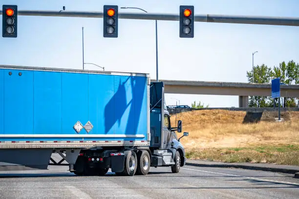 Photo of Gray day cab big rig semi truck with roof spoiler transporting cargo in dry van semi trailer turning on the crossroad intersection with traffic lights and overpass road