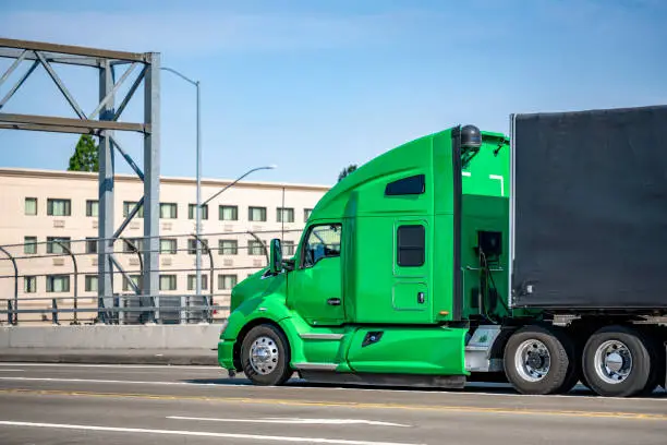 Photo of Green professional big rig bonnet semi truck transporting cargo in covered black semi trailer running on the city street bridge