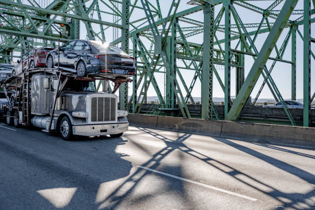 Industrial car hauler big rig gray semi truck transporting cars on modular semi trailer running on the truss Interstate Columbia River Bridge Industrial grade car hauler big rig gray semi truck transporting cars on modular two level hydraulic semi trailer running on the truss arched Interstate Columbia River Drawbridge at sunny day transporter stock pictures, royalty-free photos & images