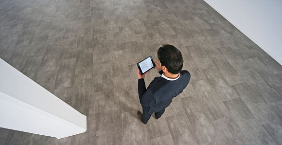 Elevated view of businessman examining blueprint while using digital tablet in unfurnished office.