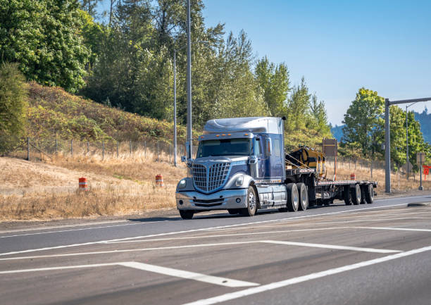 istock Stylish blue big rig bonnet star semi truck transporting construction equipment on step down semi trailer running on the highway road entrance 1343100910