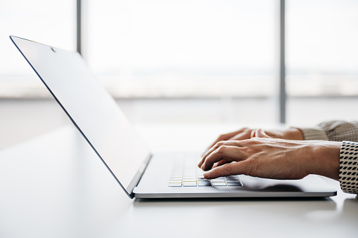 A close-up shot of a woman working on her laptop in a bright office. Her face is not visible. Horizontal daylight indoor photo with copy space.