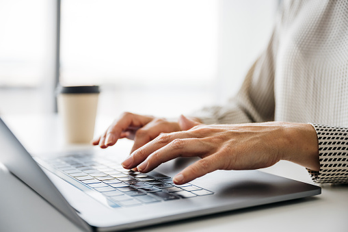 A close-up shot of a business woman's hands typing on a laptop. Her face is not visible. There is a paper coffee cup in the background. Horizontal daylight indoor photo with copy space.