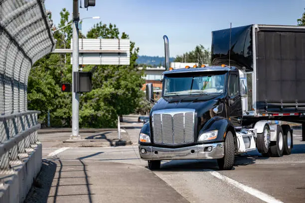 Photo of Black day cab big rig semi truck with covered dry van semi trailer with front wall spoiler turning on the city street bridge with crossroad and traffic light