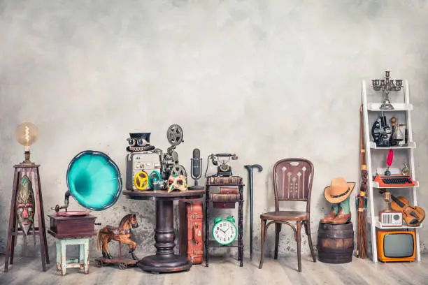Photo of Antique gramophone, chair, old typewriter, retro radio, tape recorder, projector, books, clock, camera, fiddle, mask, cylinder hat, cowboy boots, bow, cane, suitcase. Vintage style filtered photo