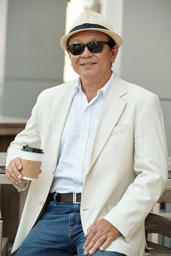 Portrait of smiling relaxed senior Vietnamese businessman in sunglasses sitting in street cafe and drinking coffee outdoors