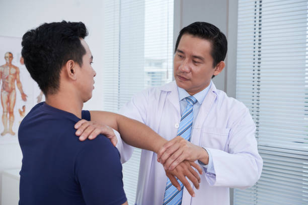 Physician Examining Sportsman Horizontal portrait of mature Asian traumatologist wearing white coat examining sporty young man palpating his knee sports medicine stock pictures, royalty-free photos & images
