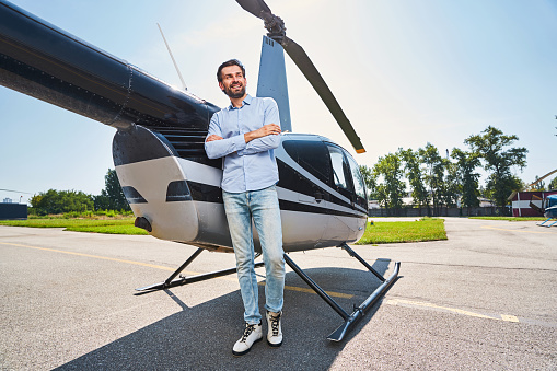 Full-length portrait of smiling confident young male leaned against private helicopter looking away