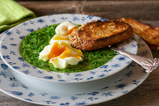 Homemade fresh cooked spinach soup served with poached eggs and roasted sunflower seed bread on rustic and wooden table. Closeup and isolated view