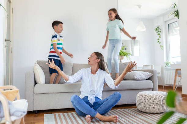 istock Tired young mother sitting on the floor and working with laptop and documents while little kids and jumping on the sofa and having fun and making noise 1343144353