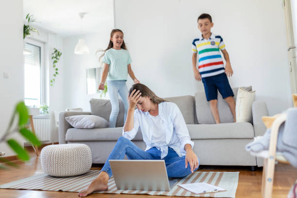 istock Tired young mother sitting on the floor and working with laptop and documents while little kids and jumping on the sofa and having fun and making noise 1343145435