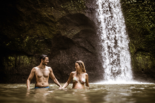 Happy couple holding hands and walking in water near waterfall. Copy space.
