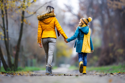 Rear view of mother and daughter talking while holding hands and walking in autumn day.