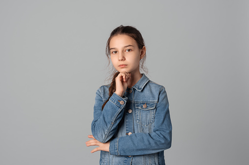 Portrait of serious preteen girl with hand on chin looking at the camera with thoughtful facial expression, isolated on grey background, wearing in denim jacket. Doubt concept