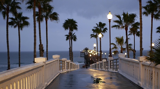 Rain drops, evening dark sky with clouds, Oceanside California USA. Empty pier, puddle, palms in twilight dusk. Reflection of lantern lights, illuminated wet broadwalk. Pacific ocean beach at night.