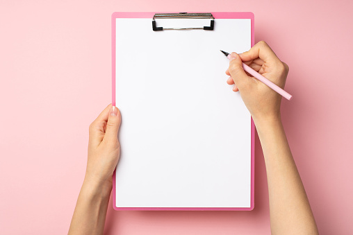 First person top view photo of female hands holding pink pen and pink clipboard with paper sheet on isolated pastel pink background with empty space
