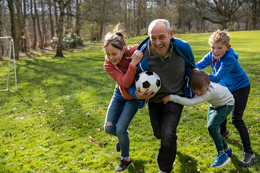 A father playing with a football with his three children on a grass area while on a staycation in Northumberland. The father is holding the football while being tackled and chased by his children.