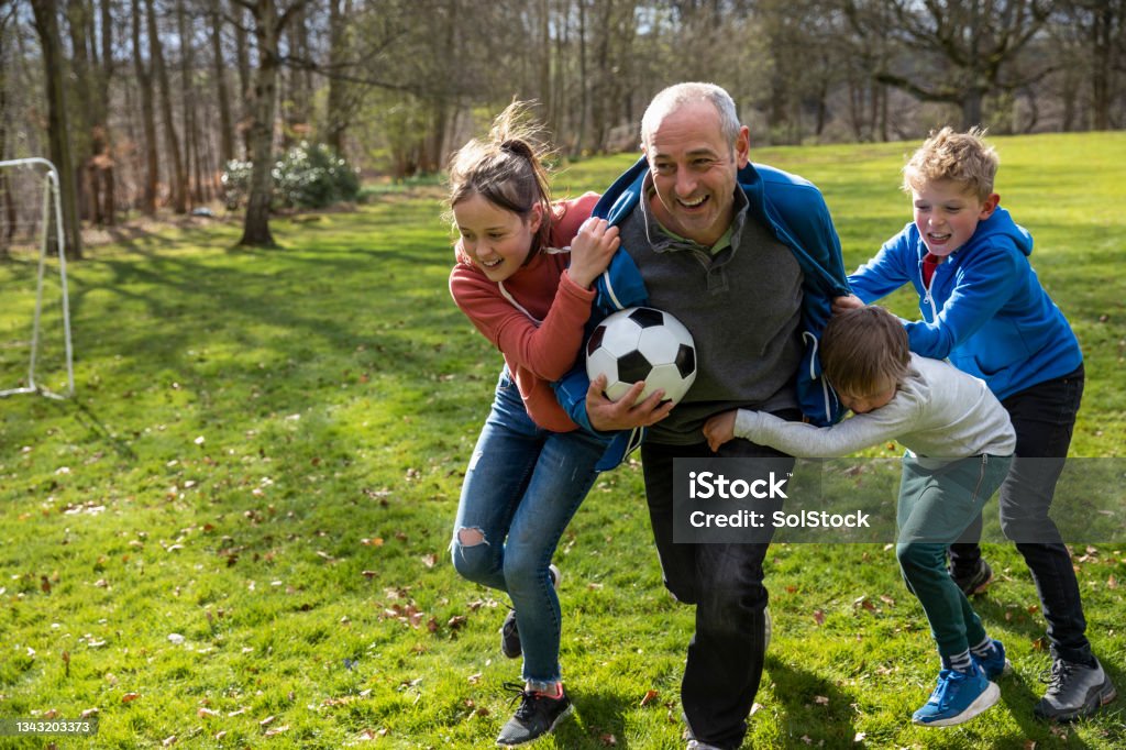 One Big Happy Family A father playing with a football with his three children on a grass area while on a staycation in Northumberland. The father is holding the football while being tackled and chased by his children. Family Stock Photo