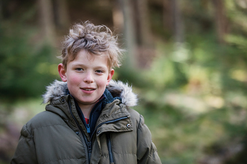A portrait of a young boy wearing a padded jacket, standing outdoors with woodland behind him. He is looking at the camera and smiling.