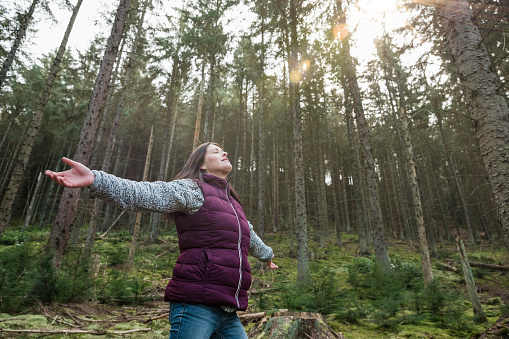 A mid adult woman standing in Thrunton Woods, Northumberland with her arms outstretched, head back and eyes closed, breathing in the fresh air from the forest, she is being at one with nature.